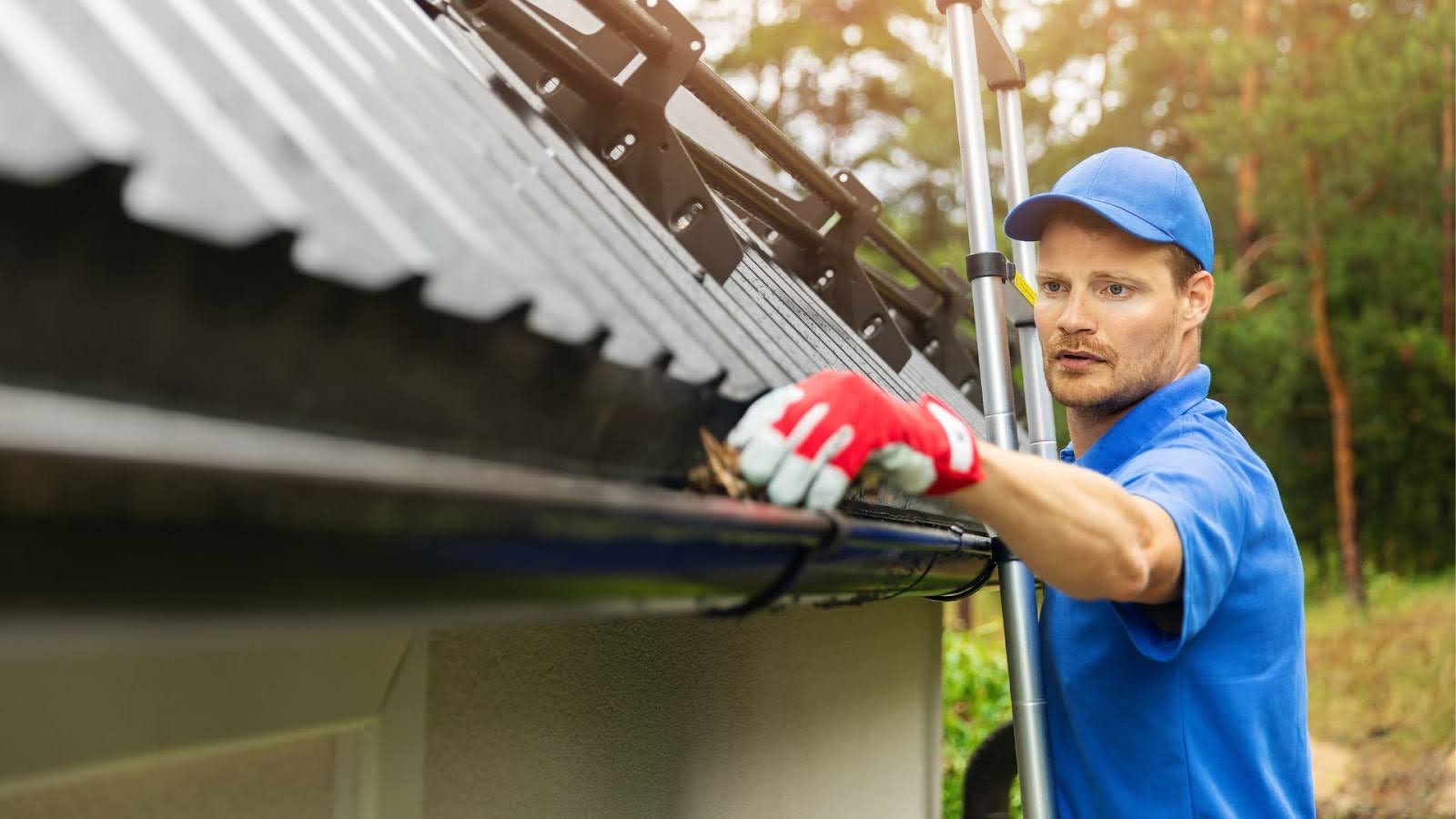 Professional gutter maintenance worker in a blue uniform manually removing leaves and debris from a house gutter, ensuring optimal function and drainage. 