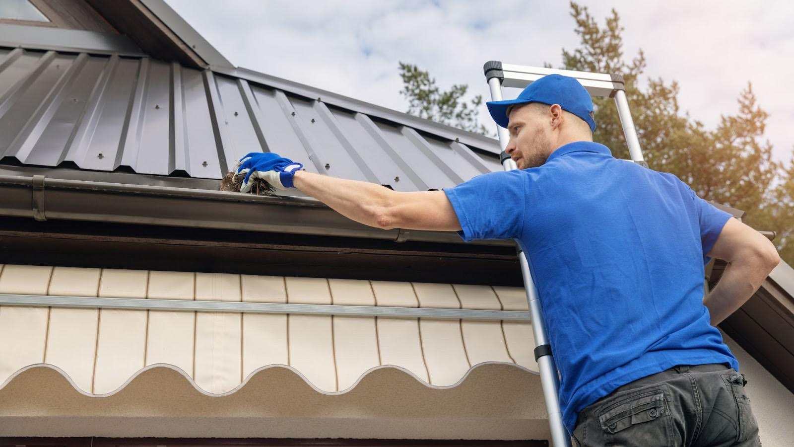 Gutter cleaning expert in blue uniform carefully removes accumulated debris by hand from a residential gutter against a backdrop of a metal roof and clear sky.