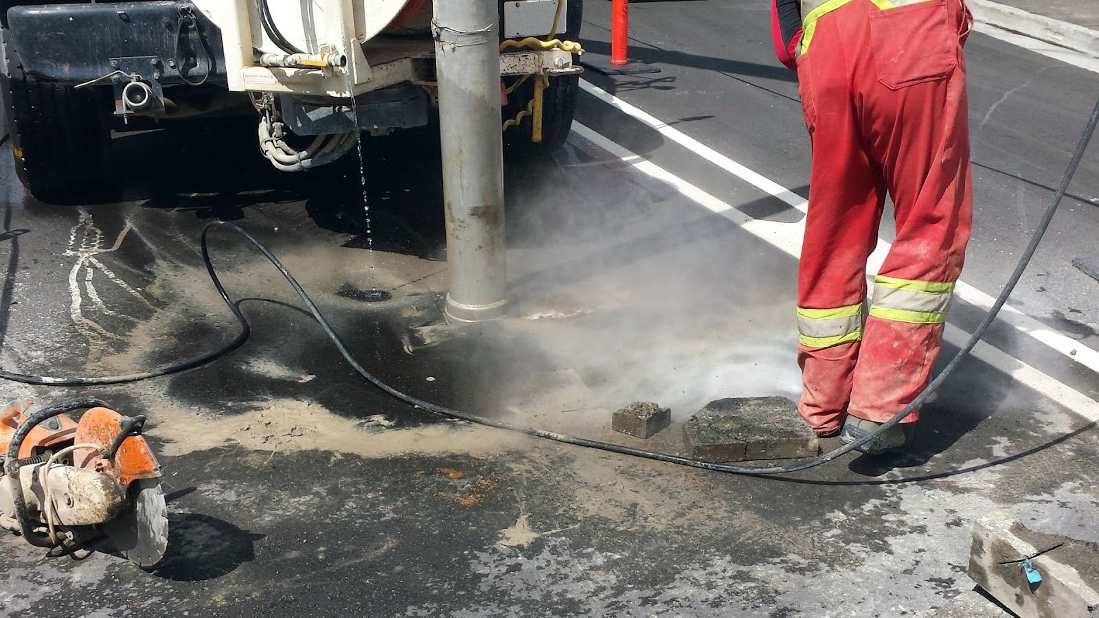 Worker in reflective safety gear conducting hydro excavation on an urban street, using high-pressure water for precision digging around underground utilities.