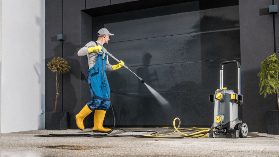 Skilled technician in blue overalls and yellow boots uses a commercial-grade power washer to clean a modern building’s exterior, demonstrating effective maintenance.