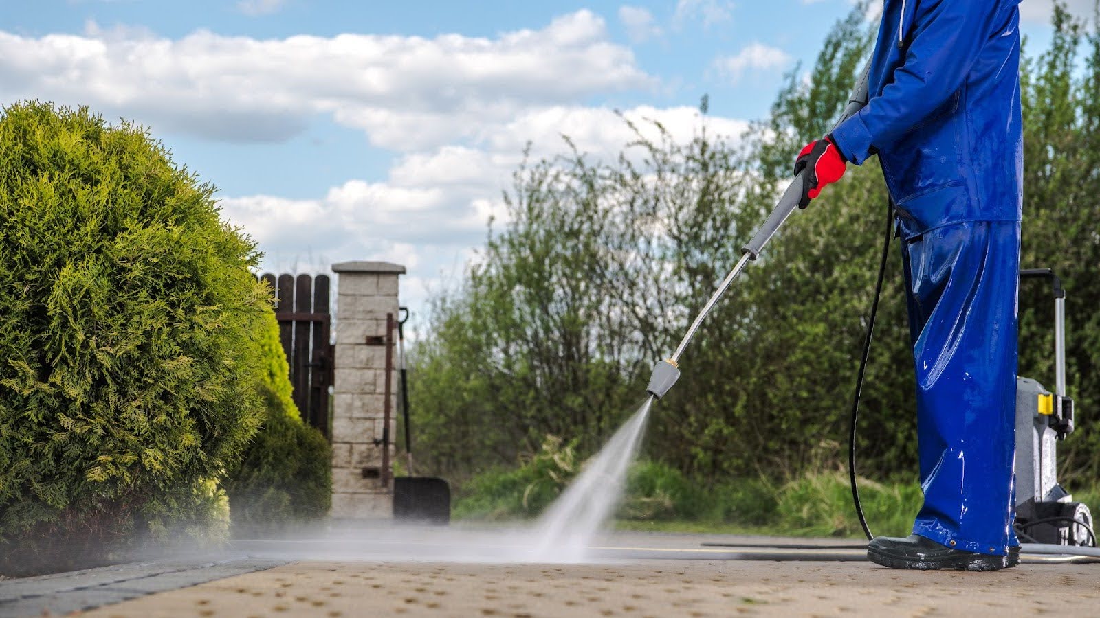 A professional cleaner in blue coveralls using a high-pressure washer on a paved area, with natural greenery in the background, demonstrating effective outdoor cleaning.