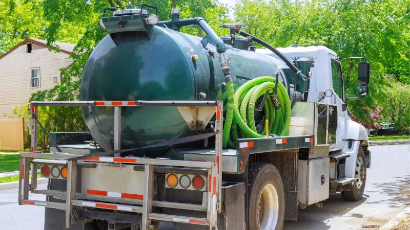 A large green vacuum truck parked on a sunny residential street, equipped with flexible green hoses for localised cleaning and waste removal services. 