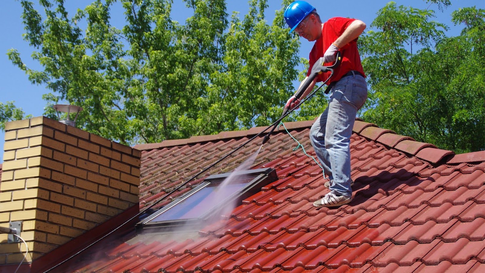 Skilled worker in a red shirt and blue safety helmet power washing a red tile roof, utilizing roof cleaning services to remove dirt and maintain the home’s structural integrity.