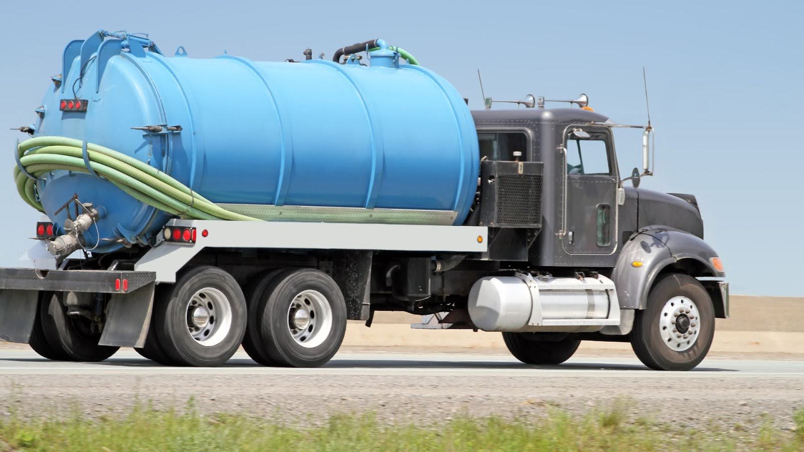 Blue vacuum truck on the move, with hoses secured on the side, driving along the highway for industrial waste management and cleaning operations. 